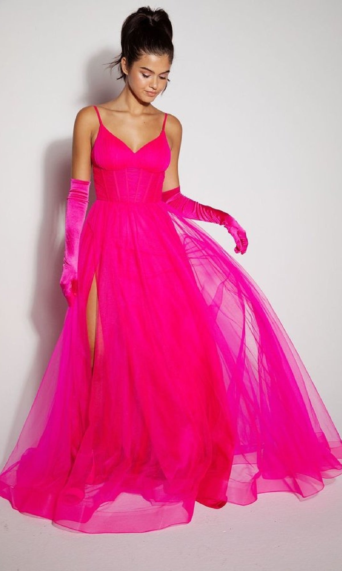 A woman poses against a plain wall in the Eureka 9177 Corset Long Tulle Prom Ball Gown, featuring a bright pink hue, sheer skirt with high slit, matching gloves, and her hair styled in a high ponytail.